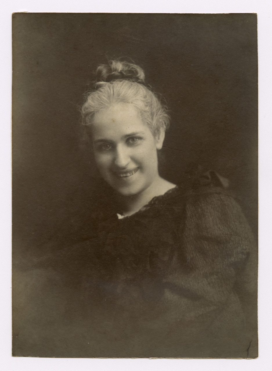 A black and white portrait of a person with an updo hairstyle and a slight smile. They are wearing a dress with a textured fabric and appear in a softly lit environment with a dark background.