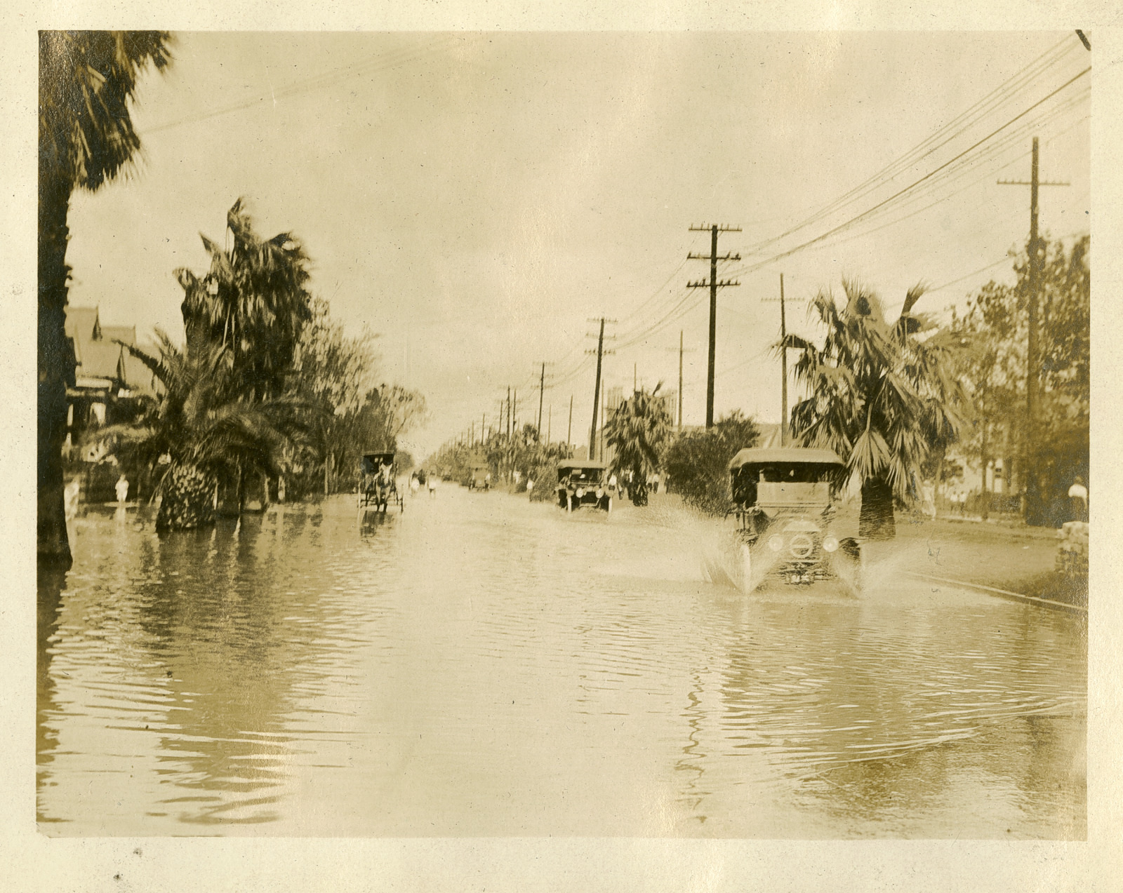 Vintage photo of a flooded street with early 20th-century cars driving through the water. Palm trees line the road, and utility poles are visible on both sides. The sky is cloudy, adding to the sepia-toned atmosphere.