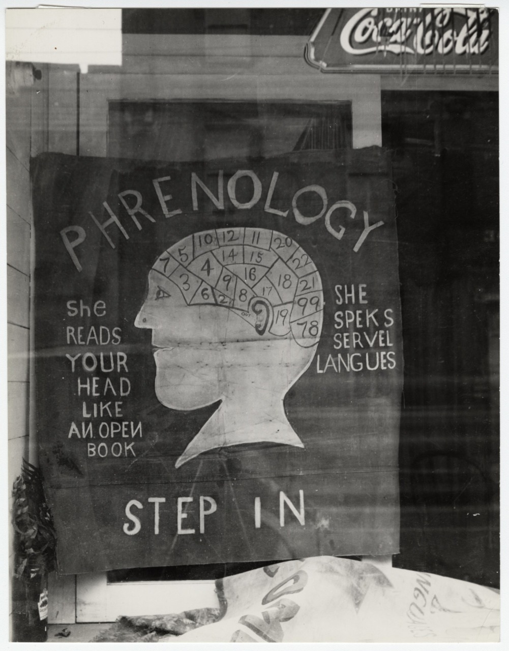 A black and white photo of a storefront display advertising phrenology. It features a human profile with numbered sections on the head, text promoting phrenology reading, and visible partial Coca-Cola signs in the background.