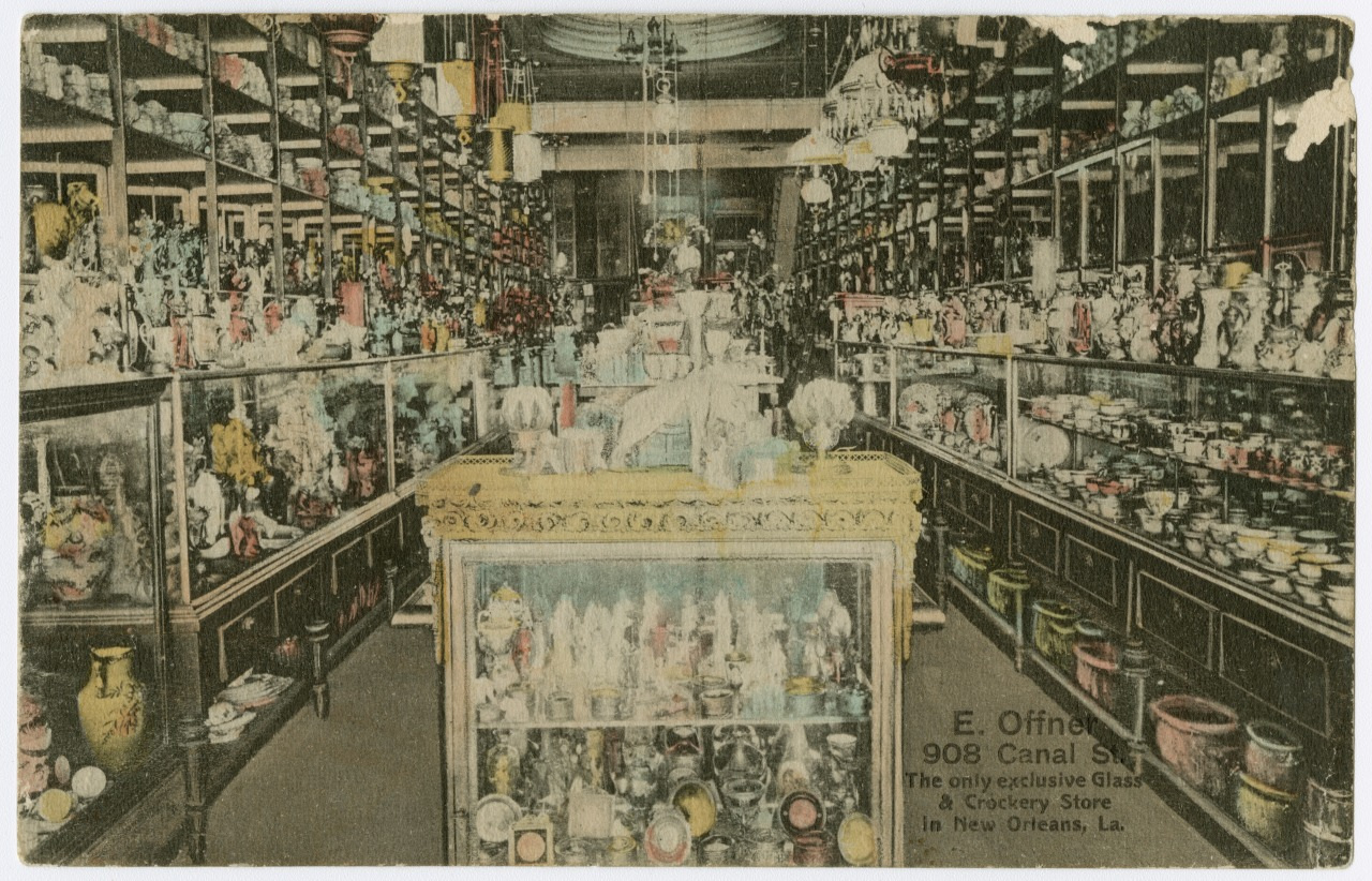 A vintage grocery store interior with ornate glass and pottery items displayed on shelves and a central glass case. The store is well-lit, and the shelves are densely packed with colorful products. Signage indicates its located in New Orleans.