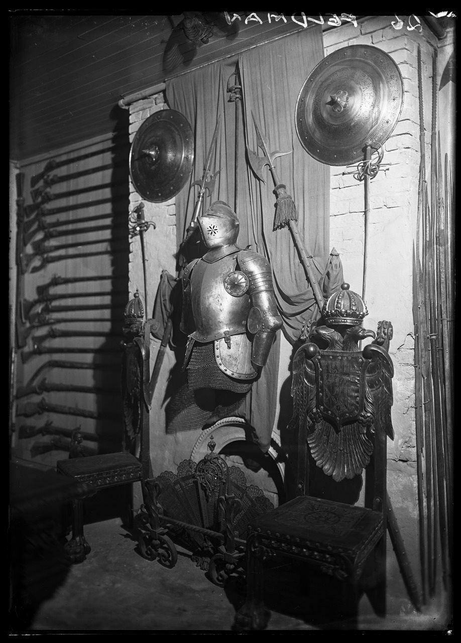 A black-and-white image featuring a collection of medieval armor and weapons. A suit of armor stands in the center with shields and spears on either side. Rifles line the wall to the left, and decorative pieces sit below the armor display.