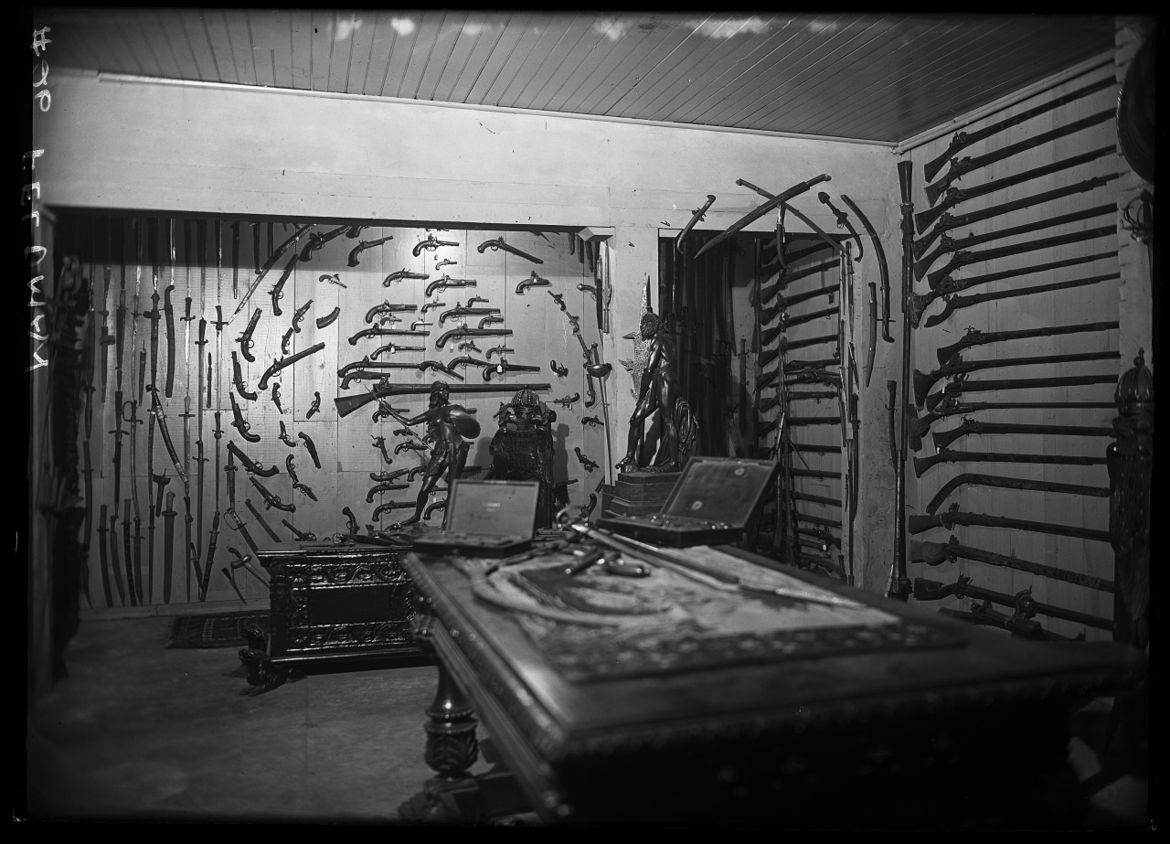 Black and white photo of a room with an extensive display of rifles and pistols on the walls. A large antique table is in the center, and there are two chairs. The room has a vintage, historical feel.