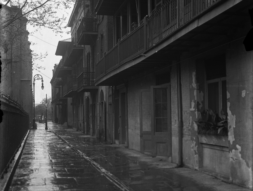 A black and white photo of a wet, empty street lined with old buildings featuring balconies. A lone figure with an umbrella walks in the distance. Leafless trees and ornate streetlamps add to the historic atmosphere.