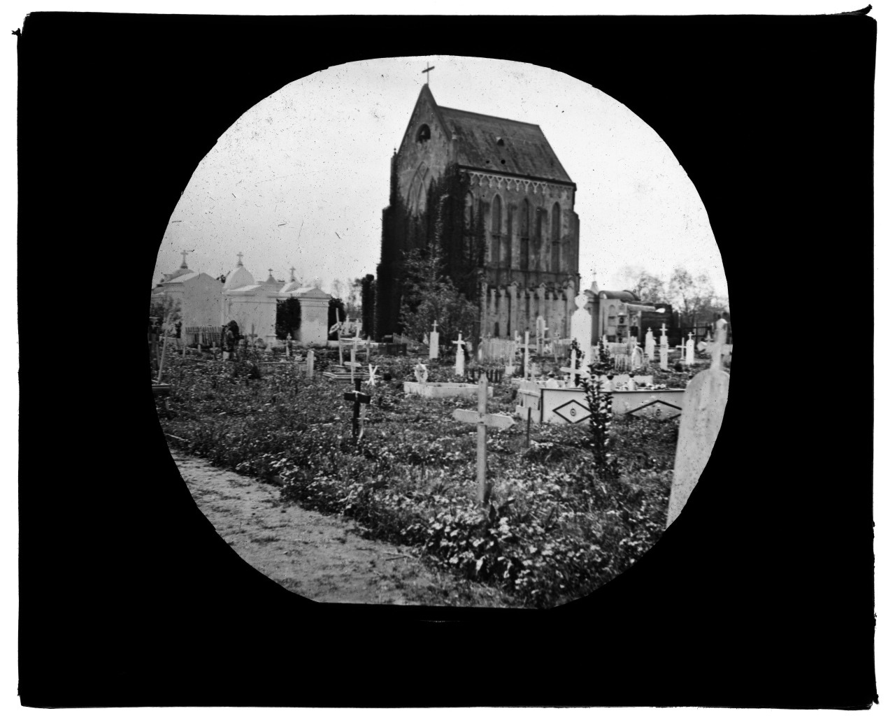 A black and white image of a cemetery with numerous cross-shaped gravestones in the foreground. A large, tall church with a steep roof and arched windows stands in the background. The ground is overgrown with grass and plants.
