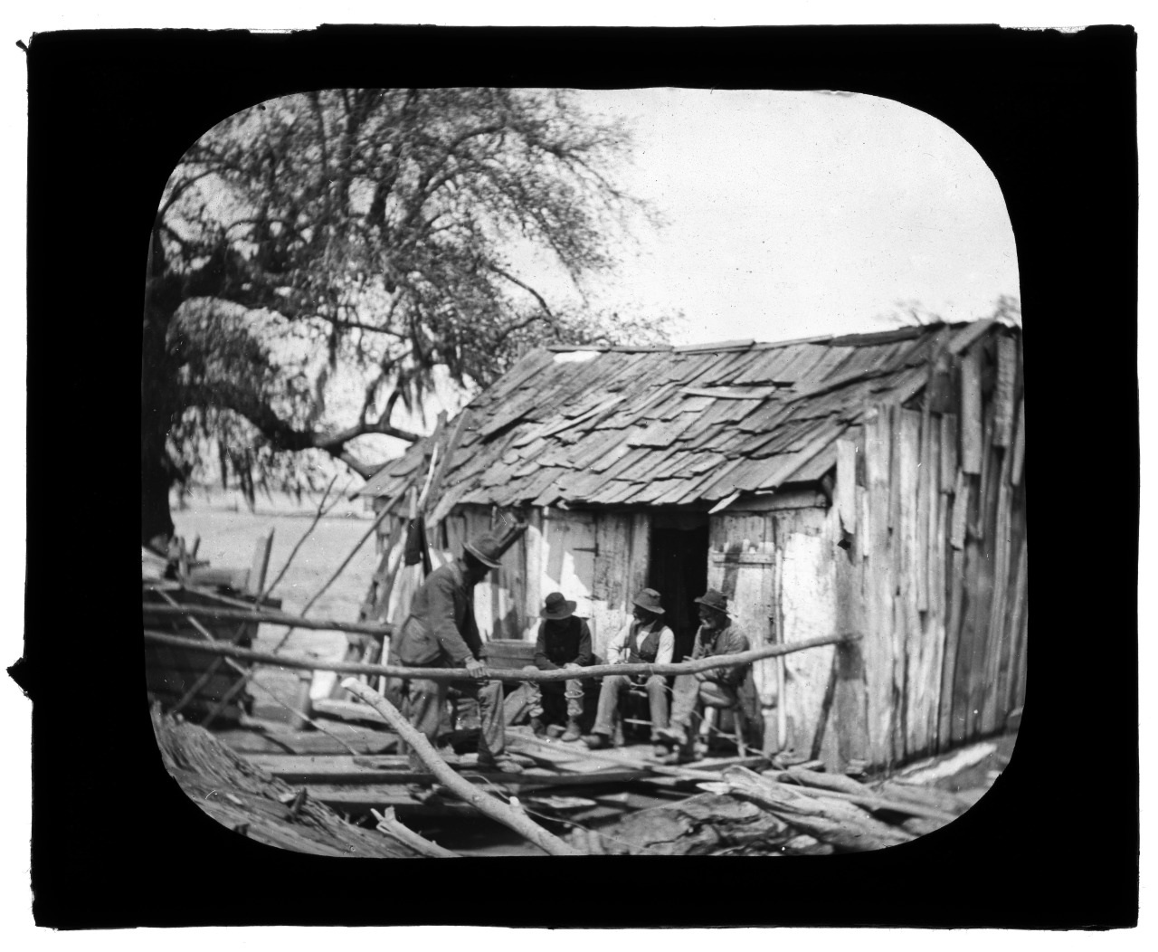 A black and white photograph of three individuals sitting in front of a weathered wooden shack with a damaged roof. Surrounding debris is visible, and a large leafless tree stands in the background. The scene conveys a rustic, historical atmosphere.