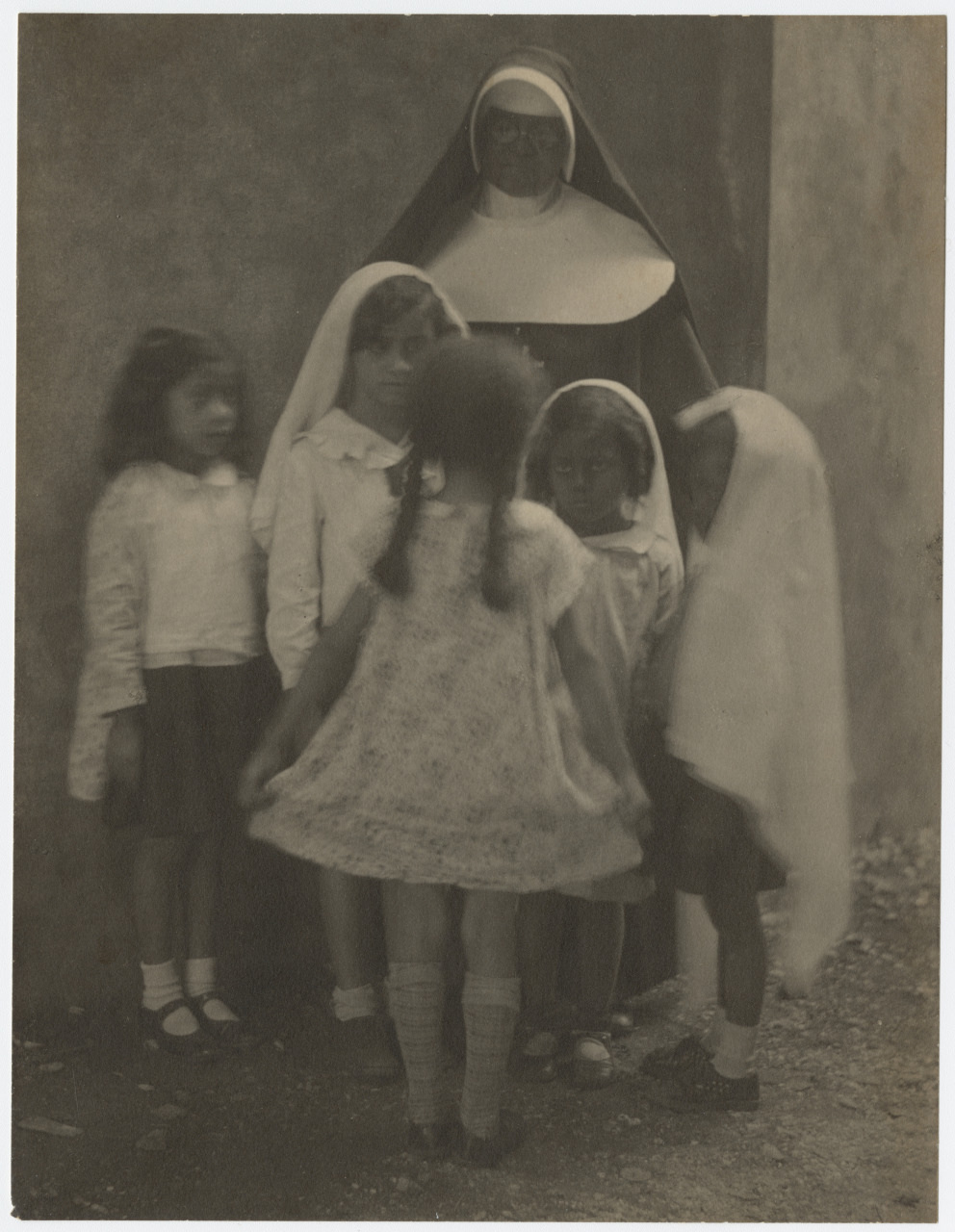 A vintage black and white photo shows a nun standing with five young girls. The children are wearing dresses, and one is standing with her back to the camera, holding out her skirt. The scene has an old-fashioned, serene atmosphere.