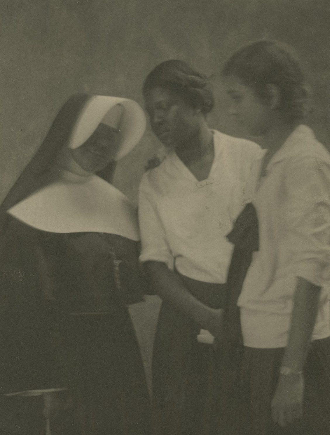 A vintage sepia-tone photograph shows a nun interacting with two adolescent girls, circa 1930.