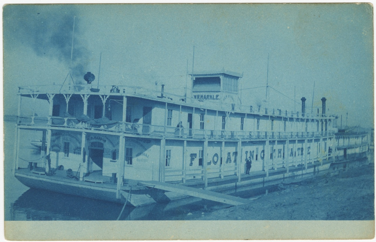 A vintage steamboat named Armadale docked at a riverbank. Signs on the side read Floating Palace. The boat has two decks and smoke billowing from its chimneys, suggesting its operational. The image has a blue tint.