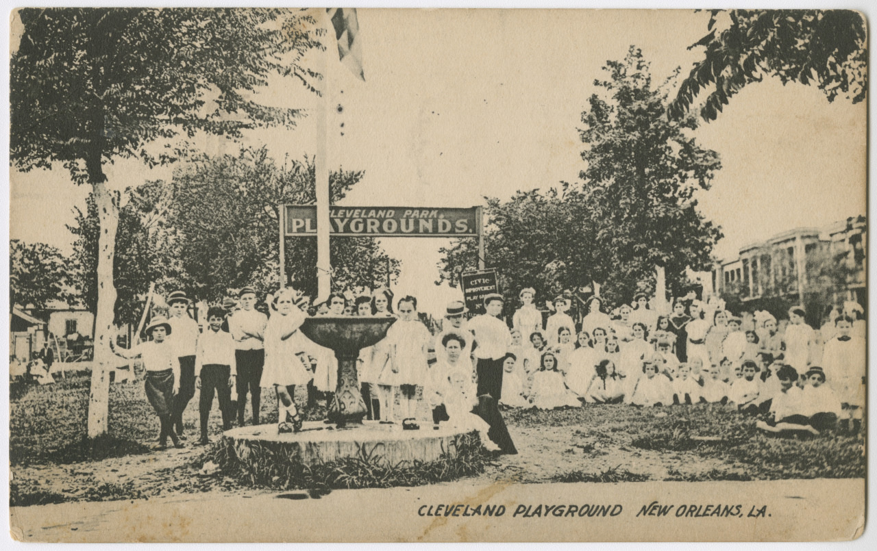 A vintage photo of a large group of children and several adults at Cleveland Playground in New Orleans, LA. They are gathered around a drinking fountain in a park setting, with trees and a sign reading Cleveland Park Playgrounds in the background.