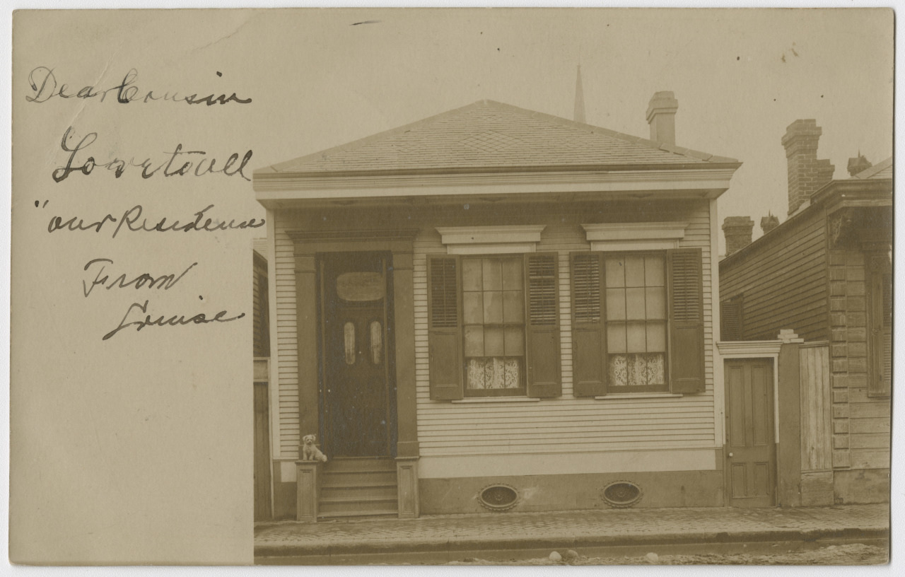 Sepia-toned photograph of a small, single-story house with a hipped roof. The house has shutters and a front door with steps. Handwritten text in cursive is visible on the left side of the image.