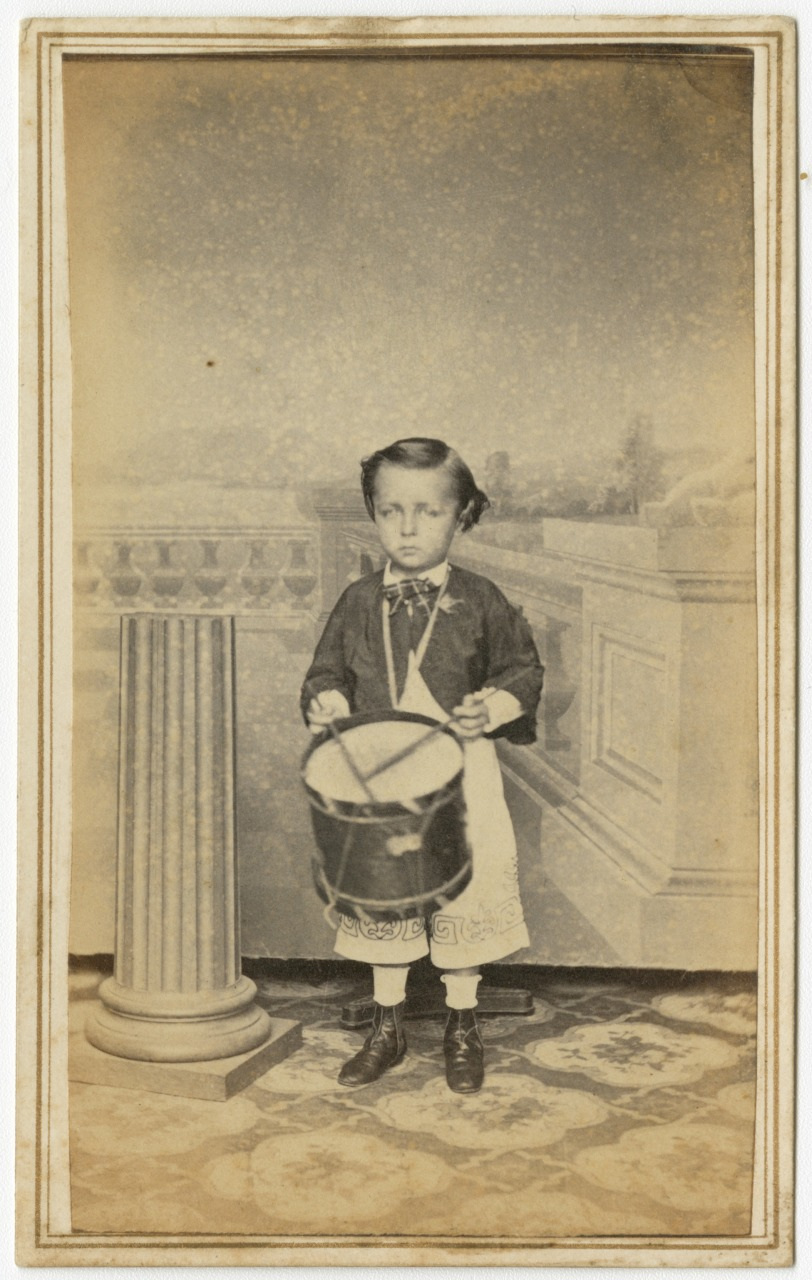 A young boy wearing a 19th-century outfit stands holding a drum in front of a decorative backdrop. The scene includes a column and ornate flooring, suggesting a formal studio setting. The boy looks directly at the camera.