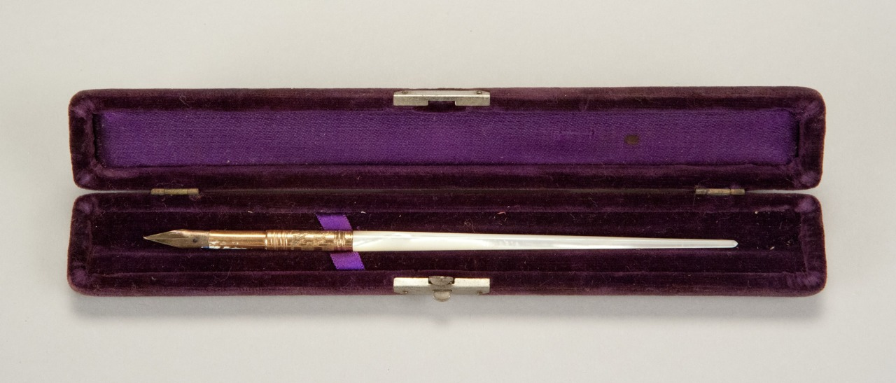 A silver dip pen with a gold ornate grip is displayed inside an open, deep purple velvet-lined case. The case is rectangular with metal clasps.