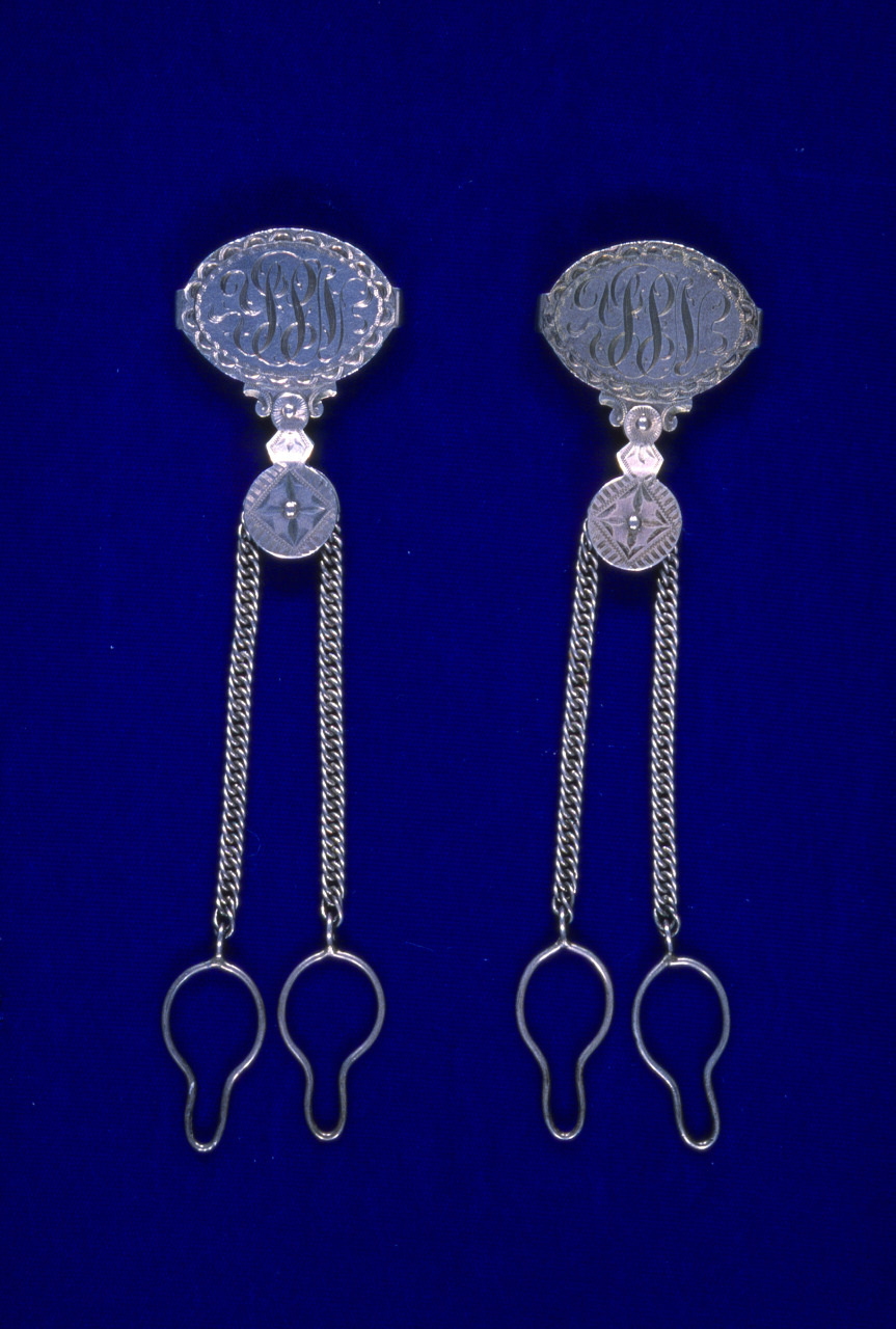 A pair of ornate silver belt clasps on a blue background. Each clasp features intricate designs and is connected to two dangling chains ending in hooks, likely used for securing or decoration.