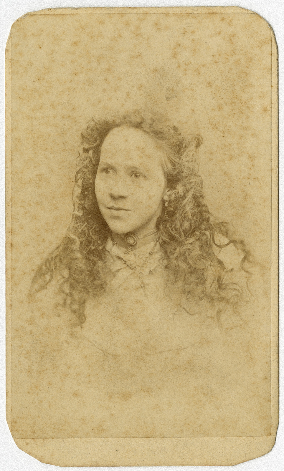 Sepia-toned vintage portrait of a young woman with long curly hair, looking to the side. She is wearing a collared garment and a necklace, with a neutral expression, set against a plain background. The image shows signs of aging and wear.