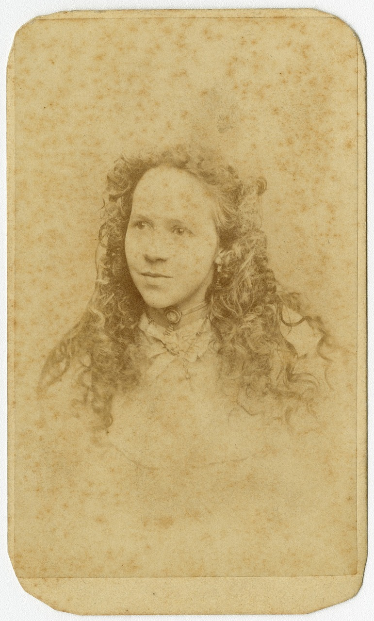 Sepia-toned vintage photograph of a young woman with long, curly hair. She gazes slightly to the left, wearing a choker and a light-colored dress. The background is plain, and the image has a worn, aged appearance.
