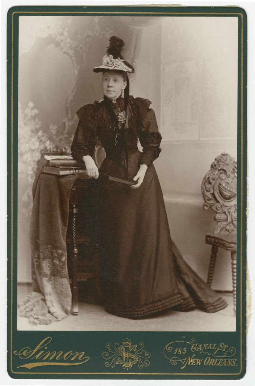 A woman in a Victorian-era dress stands beside a small round table with books. She wears a hat adorned with feathers and holds a fan. The backdrop is ornate with floral details and a decorative chair is partially visible.
