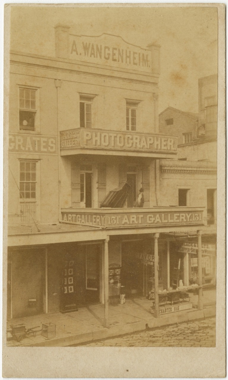 A sepia-toned photo of a multi-story building with signs for A. Wangenheim and Photographer. The building features balconies and a sidewalk storefront with various signs and displays. People are visible on the balconies and sidewalk.