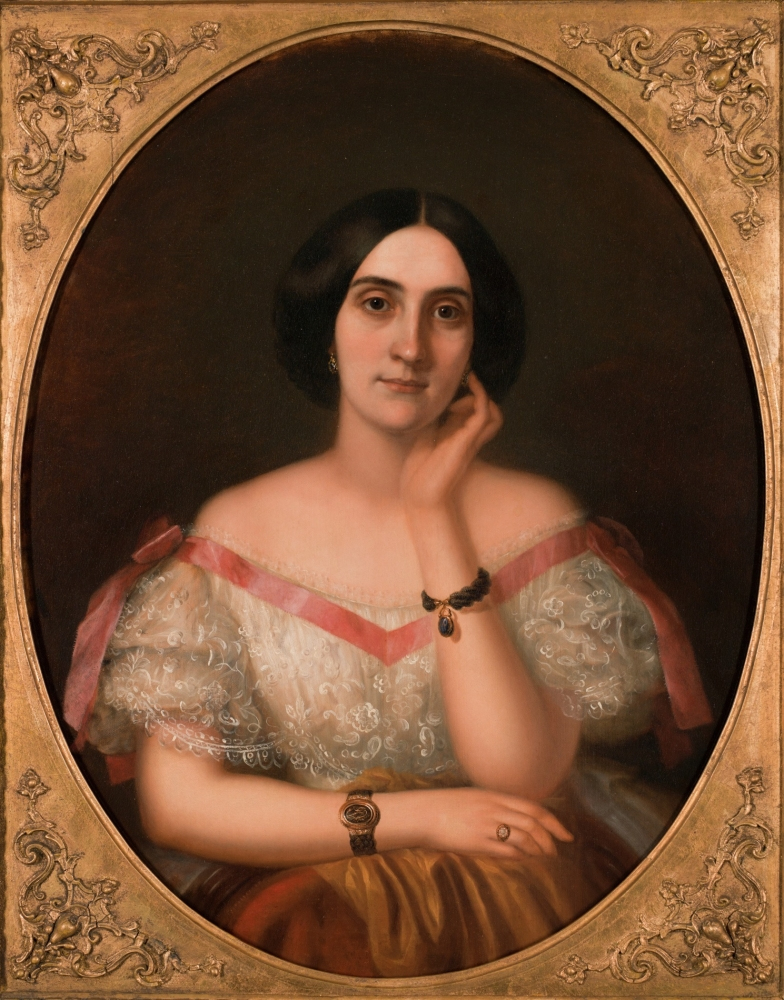 A vintage portrait of a woman in a lace-trimmed dress with pink ribbon accents. She has dark hair and wears a bracelet and earrings. Her hand rests on her cheek, and she sits against a dark background within an ornate frame.
