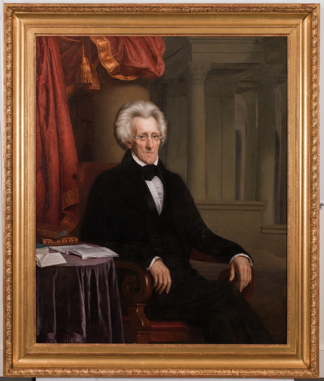 A portrait of an elderly man with white hair, wearing glasses and a black suit, seated in a red chair. He rests his arm on a table with papers under a red curtain, and a grand architectural backdrop. Framed in gold.