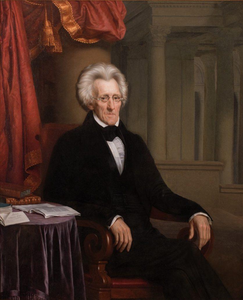 A portrait of an elderly man with white hair wearing a dark suit and glasses, seated in an ornate chair. A red curtain and architectural columns are in the background, with papers on a table beside him.