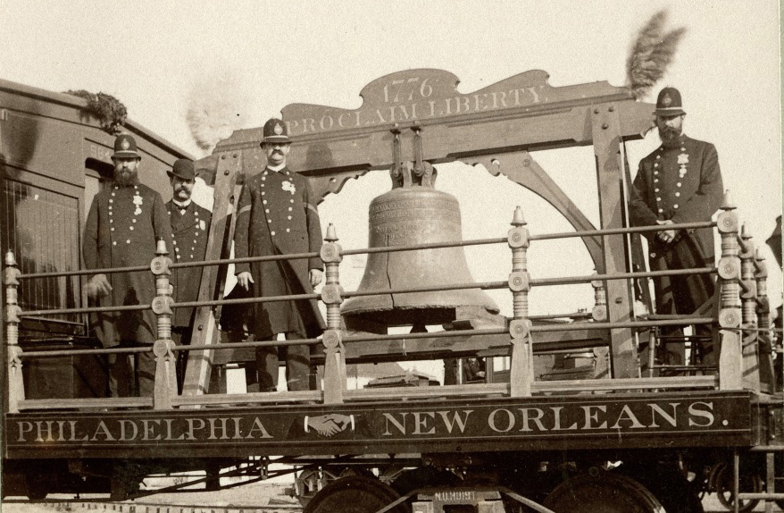 A historic photograph depicts four uniformed men standing beside the Liberty Bell, which is mounted on a railcar platform. The platform is labeled Philadelphia and New Orleans, showing the bells transport.