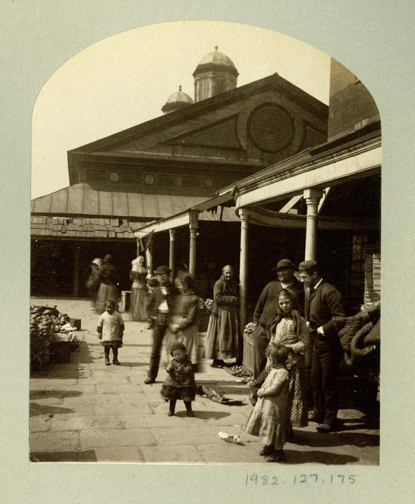 A historical scene shows people in period clothing gathered outside a building with a dome. A man and woman stand by baskets, children play, and others walk nearby, with a covered walkway and market stalls in the background.
