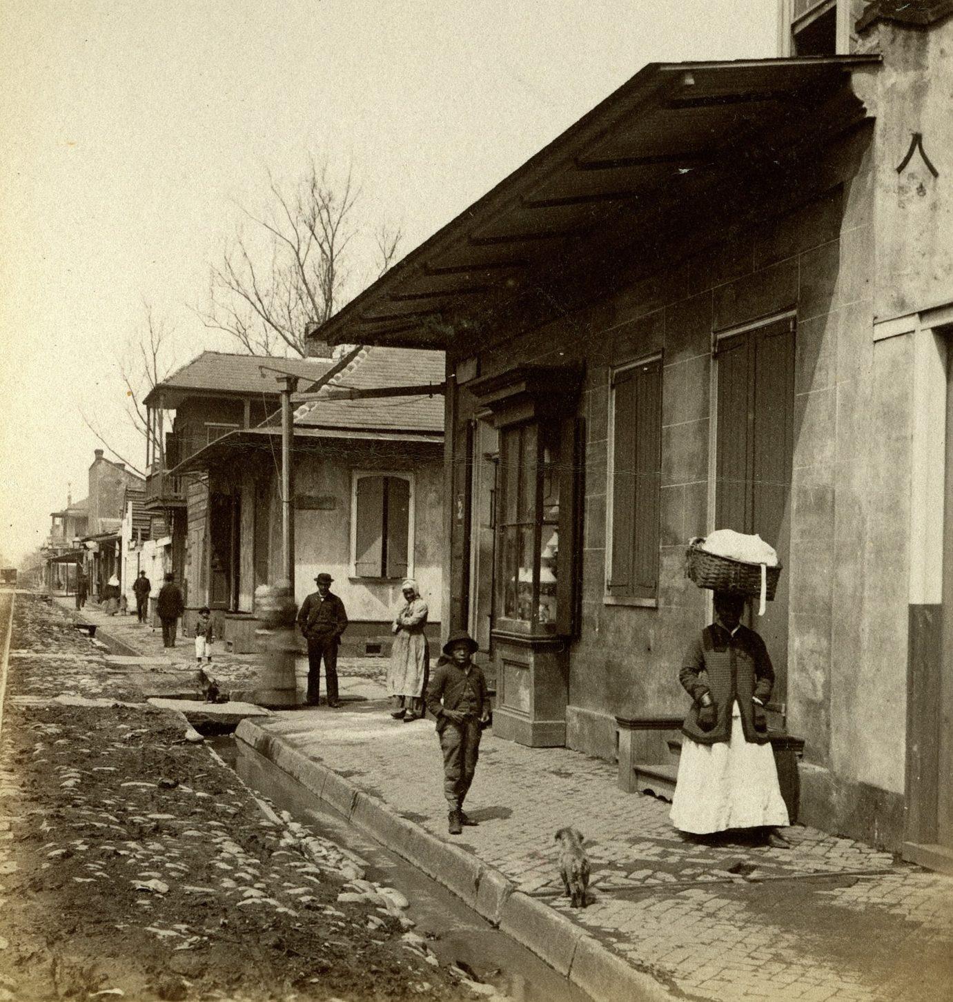 A vintage street scene near a railway line shows a woman carrying a basket on her head, a child walking, and several adults standing. Buildings line the street, with trees and a cloudy sky in the background.