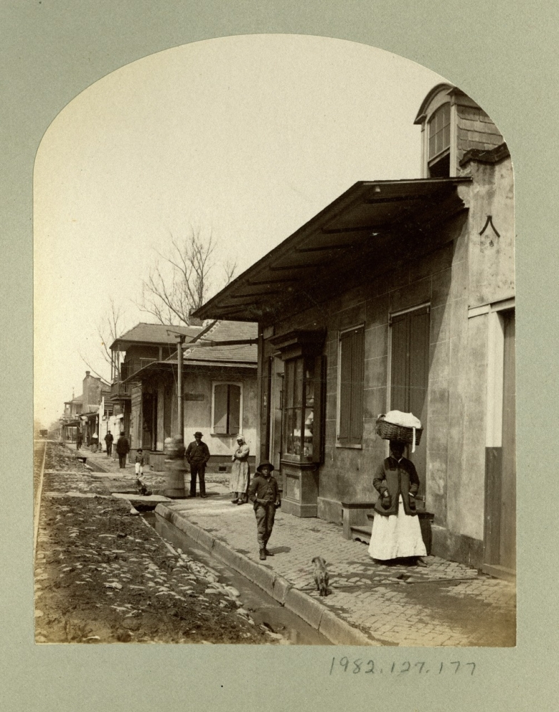 A vintage photograph shows a street scene with people standing or walking on a sidewalk beside a row of buildings. One woman holds an umbrella. The ground appears muddy, and leafless trees line the street. The image has an antique, sepia tone.