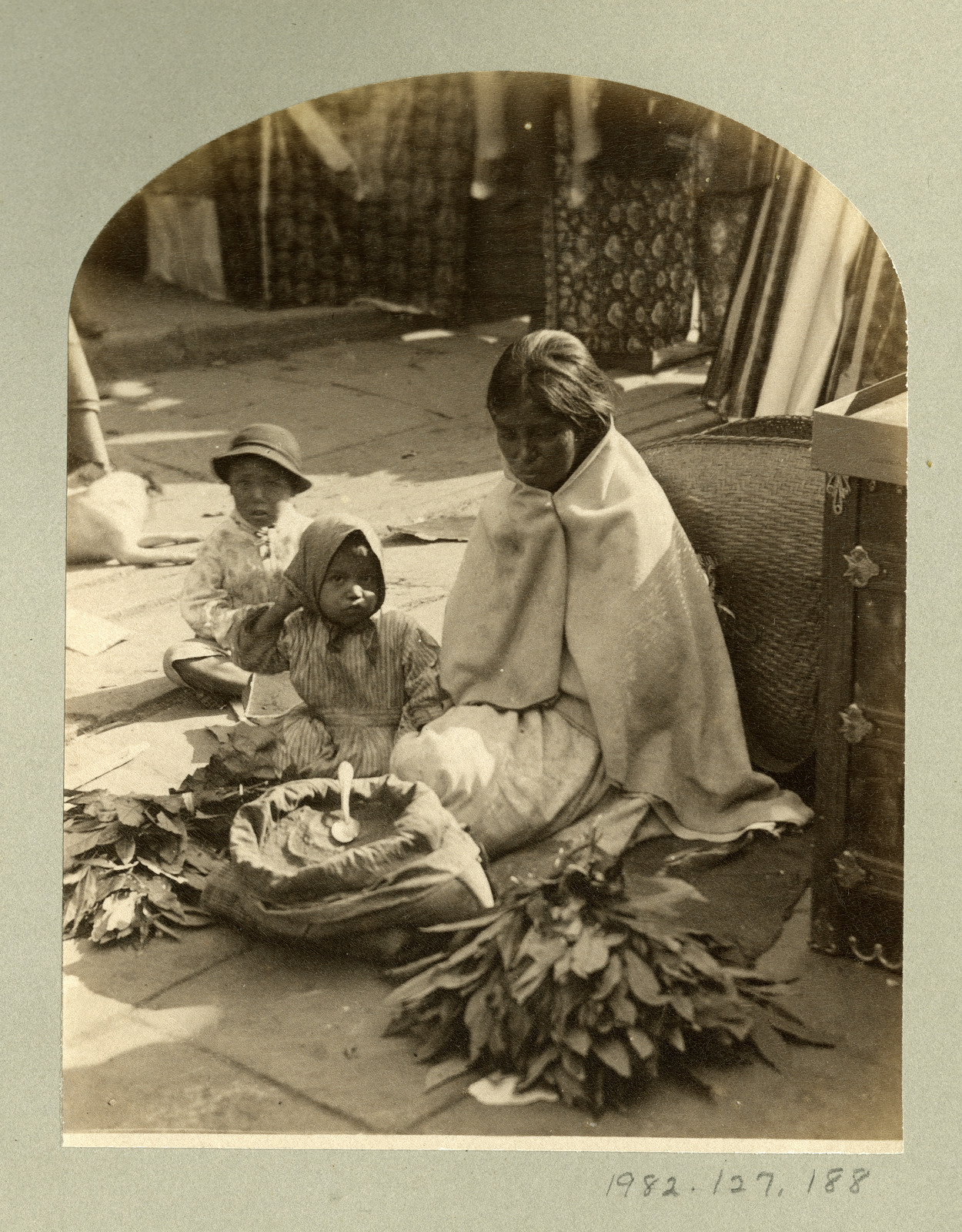 A photographic print shows an American Indian woman sitting on the ground among the stalls of the French Market in New Orleans selling file. The woman has both an open sack of file powder, and two bundles of leaves. A girl and a boy are sitting on the ground next to her.