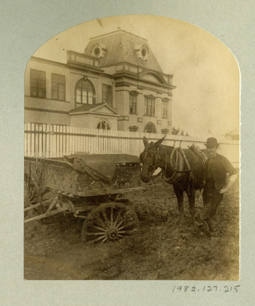 A sepia-toned old photograph depicts a man standing beside a horse-drawn cart filled with materials. A large building with arched windows and a fenced yard is in the background. The scene suggests a historical urban setting.