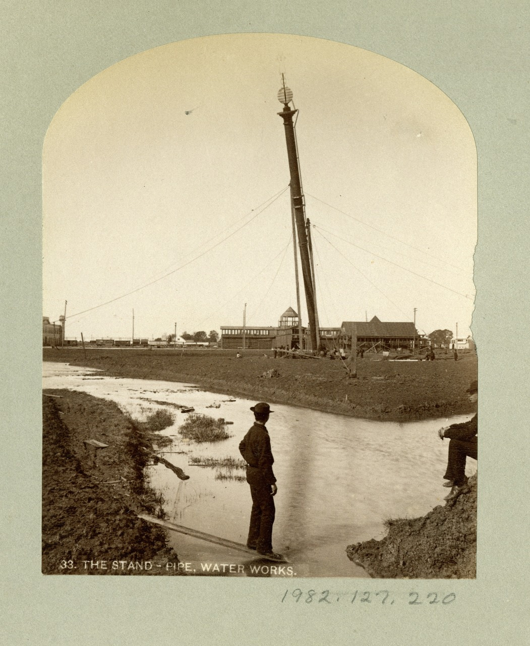 A sepia-toned photograph of a man standing by a riverbank, observing a tall, vertical structure under construction in the background. Nearby, another person sits on the bank, and several buildings are visible. The photo is labeled The Stand-Pipe, Water Works.