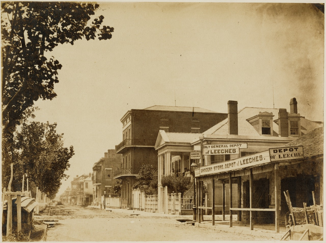 A sepia-toned photograph of a late 19th-century street scene. Buildings line both sides of a dirt road. A sign reads Grocery Store Depot W. Leche. Leafy trees border the sidewalk, and the area appears quiet and deserted.