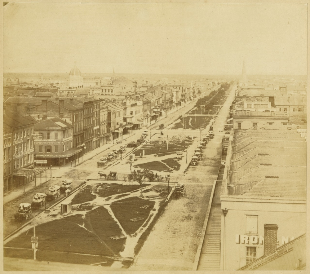 A sepia-toned aerial view of a wide street lined with horse-drawn carriages, surrounded by historic buildings. The street features a landscaped median with pathways and sparse vegetation, extending toward the horizon.