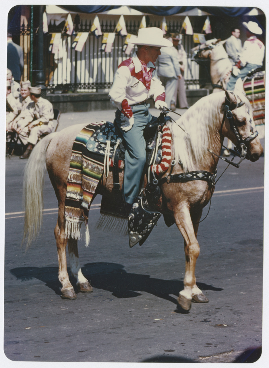 A person dressed in a cowboy hat, red scarf, and gloves rides a horse adorned with decorative blankets during a parade. People in matching outfits watch from the sidewalk.