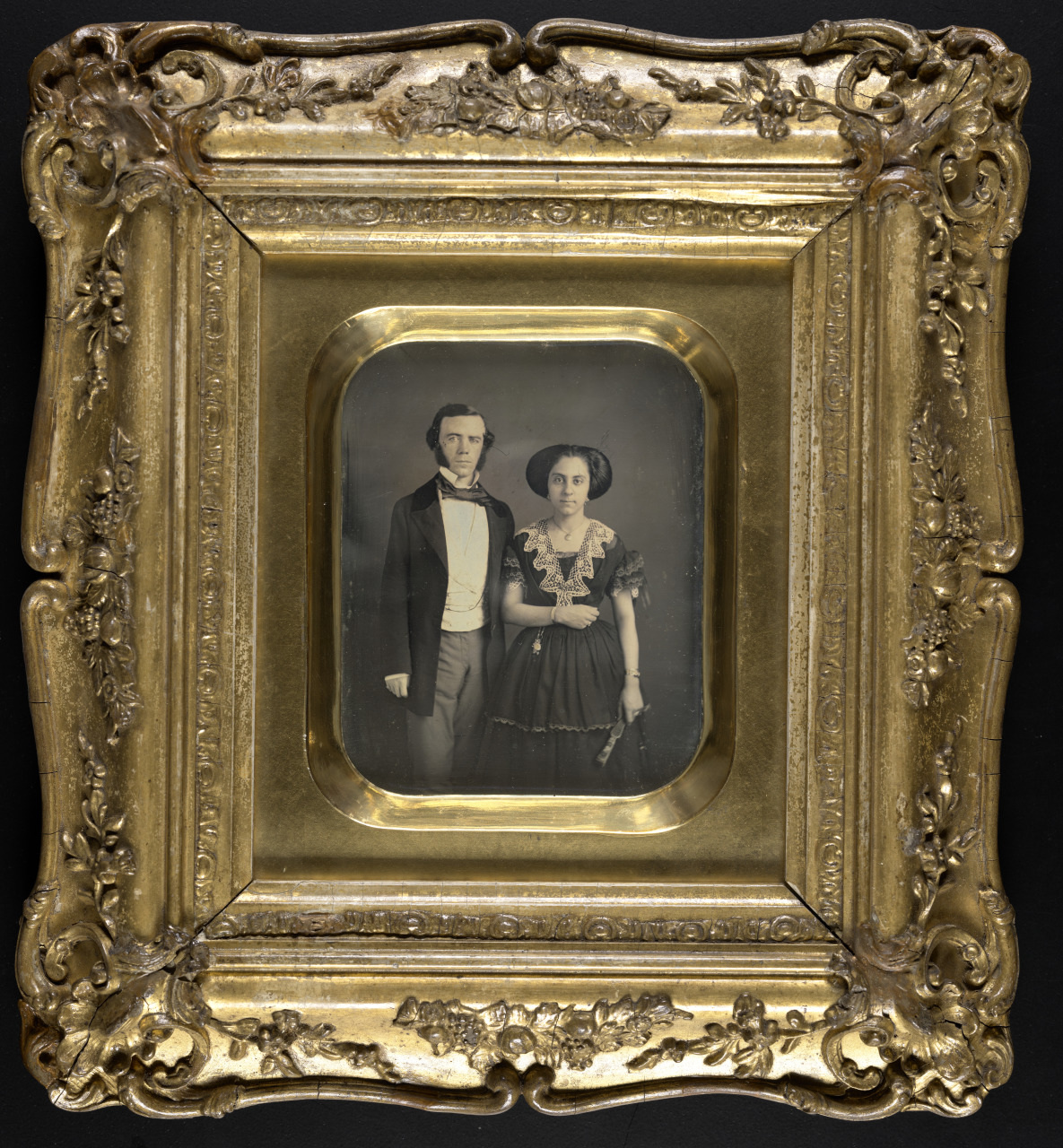 A vintage portrait in an ornate gold frame features a man and woman in formal 19th-century attire. The man wears a dark suit, while the woman is in a dark dress with lace details, holding a small fan.