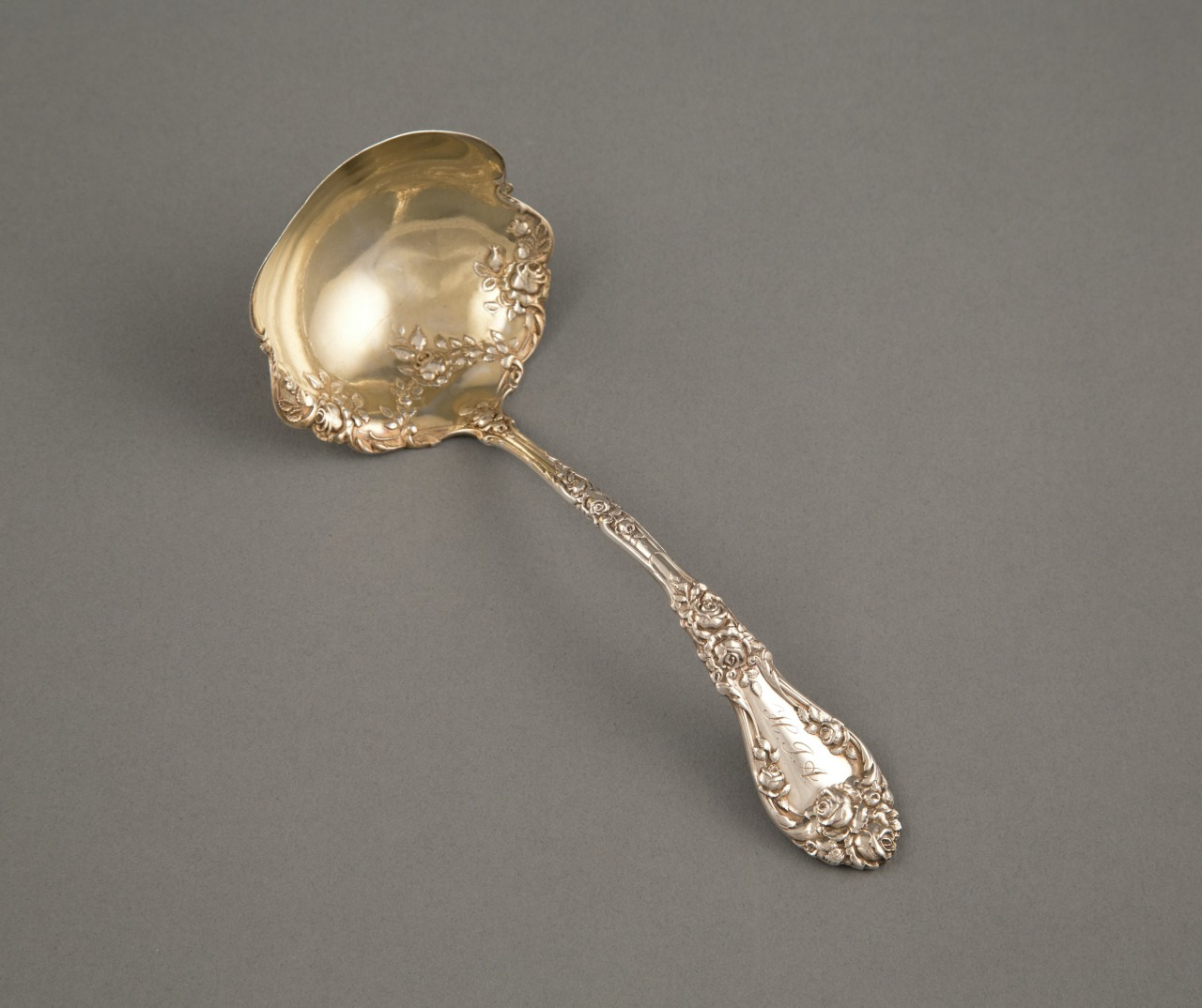 An ornate silver ladle with an intricately engraved handle and a polished, deep bowl, lying on a plain gray background.