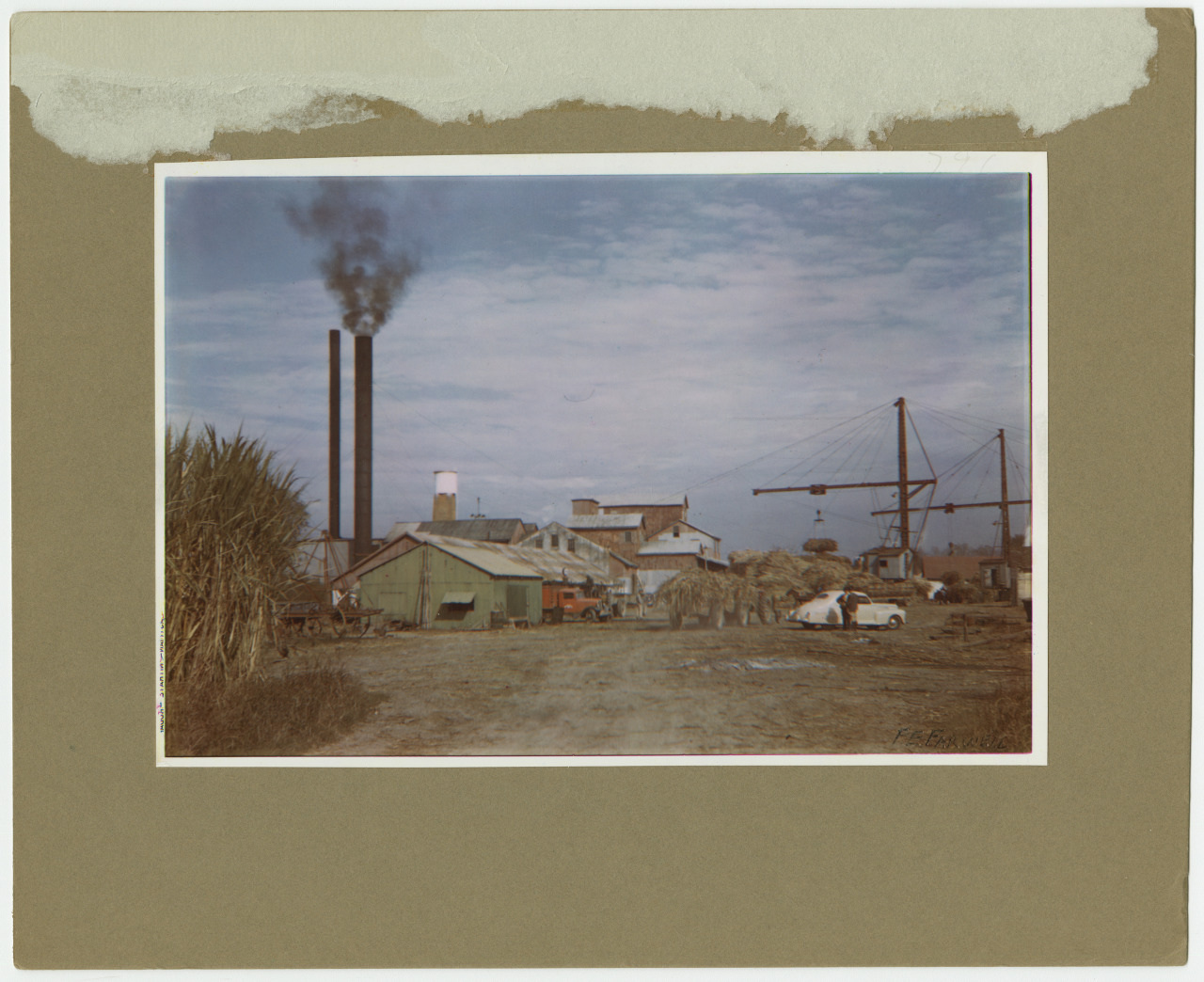 A vintage photograph of an industrial site with two tall smokestacks emitting smoke. There are buildings, a crane, and piles of material. The foreground has a dirt path and some greenery on the left, with a partly cloudy sky above.