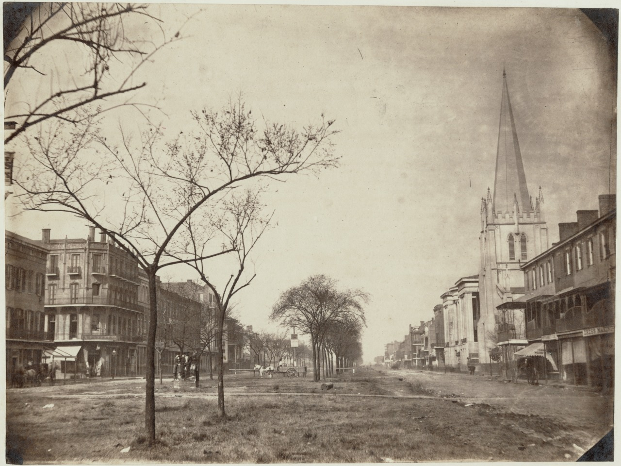 A historic black and white photo of a wide street lined with trees and buildings. At the right, a large church with a tall spire stands prominently. People are walking along the sidewalk, and horse-drawn carriages are parked along the street.