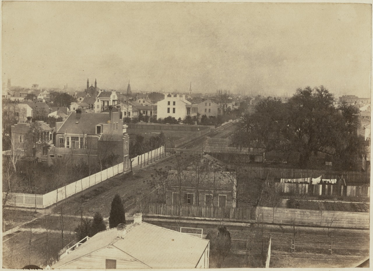 A sepia-toned photograph of a 19th-century urban neighborhood. The image shows houses with pitched roofs lining dirt roads, surrounded by wooden fences. Leafless trees and distant church spires are visible under a cloudy sky.