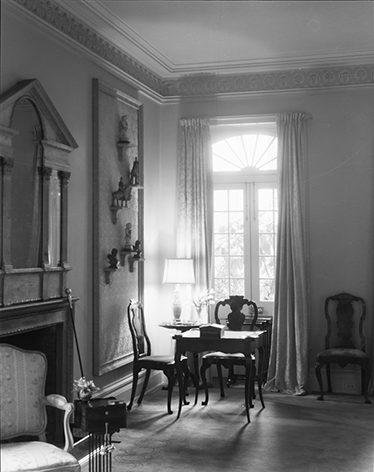 A black and white image of a classic living room featuring a large window with curtains, a wooden table with chairs, a table lamp, and ornate wall decorations. A fireplace is on the left, and a patterned ceiling adds elegance to the space.