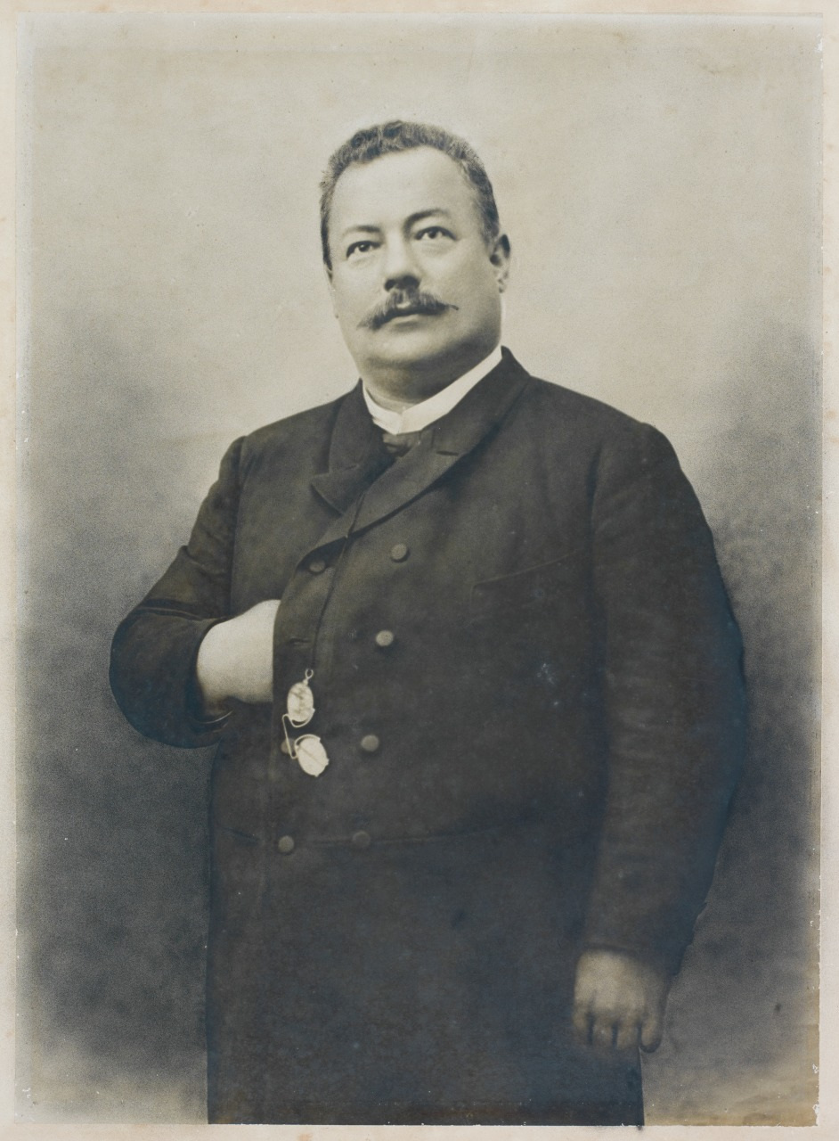A sepia-toned vintage photograph of a man with a mustache, dressed in a formal, dark suit. He has one hand tucked into his jacket and is gazing into the distance. The image conveys an early 20th-century style.
