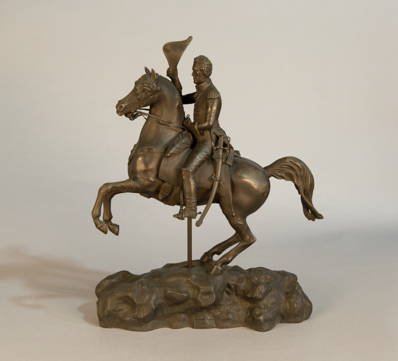 A bronze statue of a uniformed rider on a rearing horse atop a rocky base. The rider holds the reins in one hand and appears to be wearing a detailed military outfit with a hat. The sculpture is displayed against a neutral background.