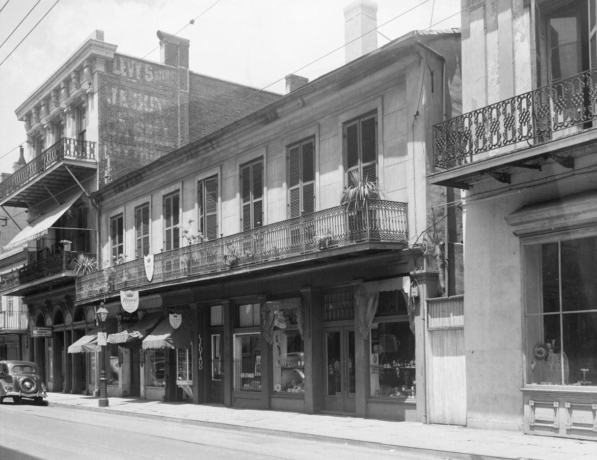 A black and white photo of a historic urban street scene featuring two-story buildings with wrought iron balconies and awnings. A vintage car is parked along the street. Signs and storefronts line the road, capturing a classic architectural style.
