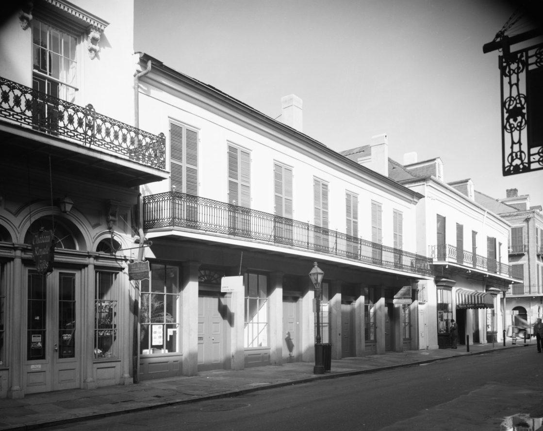 Black and white photo of a historic street scene with buildings featuring wrought iron balconies, large doors, and shuttered windows. A streetlamp is visible along the sidewalk.