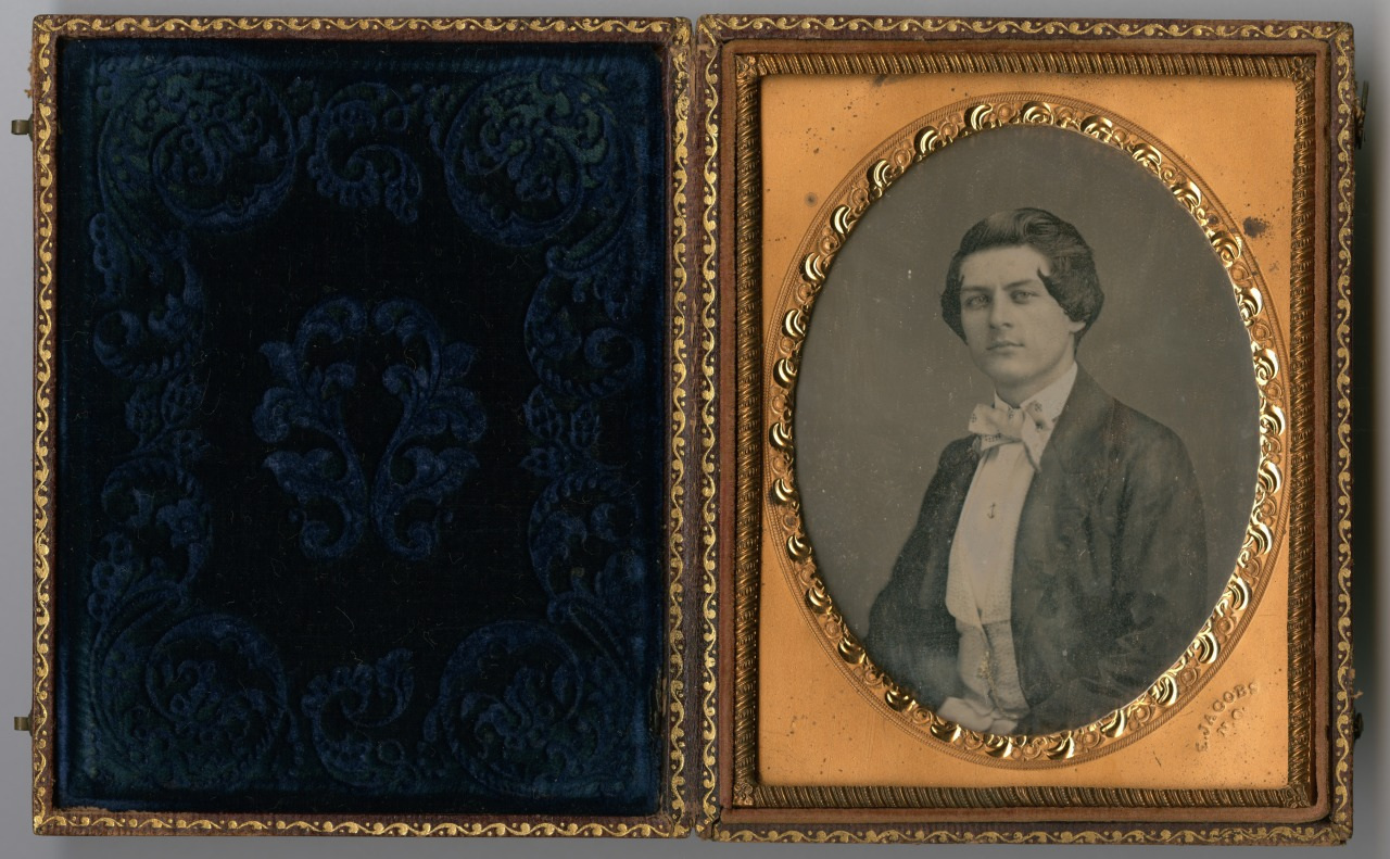 An antique sepia-toned portrait of a young man in formal attire, set in an ornate oval frame. The frame is part of a decorative case with an intricate pattern on the opposite panel.