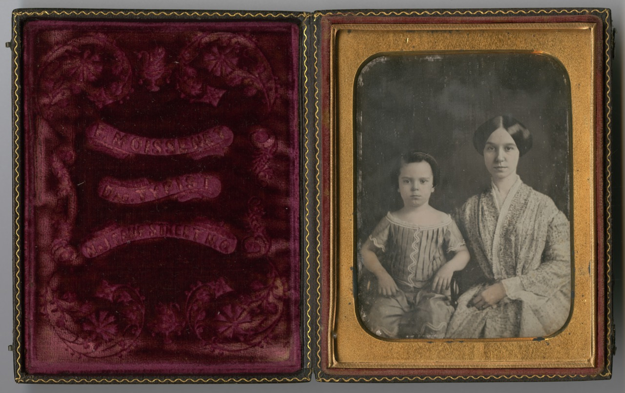 A vintage, sepia-toned portrait in an ornate frame shows a woman seated next to a young child. Both wear 19th-century clothing. The frame features intricate floral designs and sits next to a decorative velvet-like cover.