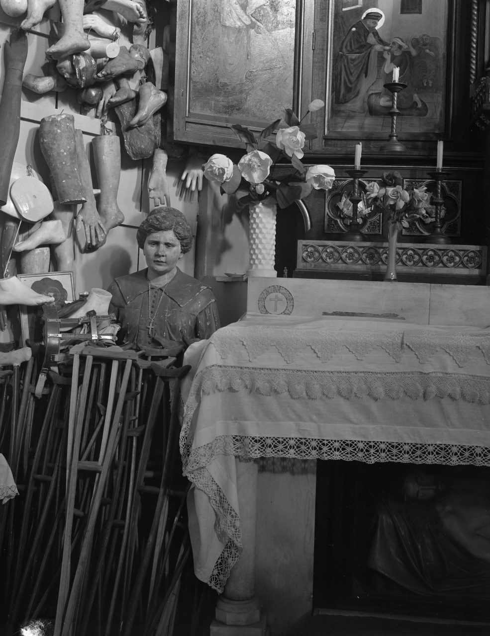 A black and white image of a shrine with mannequin limbs displayed on the walls. A mannequin dressed as a woman sits beside crutches near an altar. The altar is draped with a cloth, adorned with flowers and candles, and a religious painting is above it.
