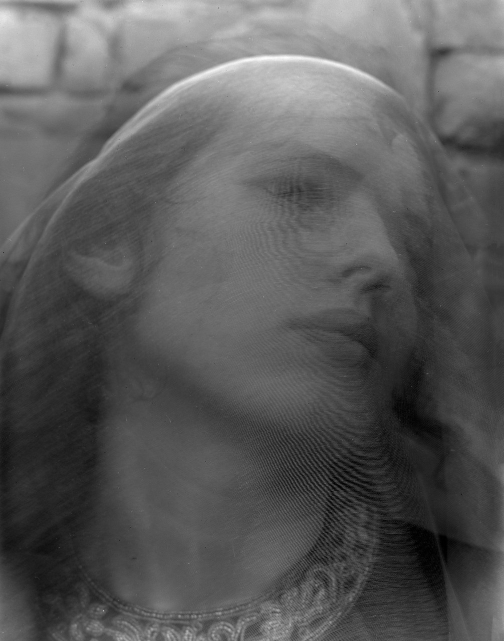 A black and white photograph shows a womans face with a serene expression. Her head is tilted slightly back and she is draped in a sheer veil. An intricate necklace adorns her neck, and a stone wall is faintly visible in the background.