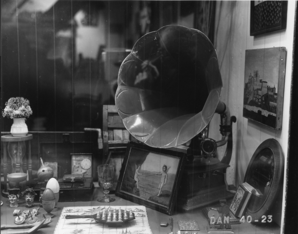 A vintage shop window display featuring a gramophone, framed artwork, a clock, ornaments, and a potted plant. The arrangement includes a mix of decorative and functional items in a nostalgic setting.