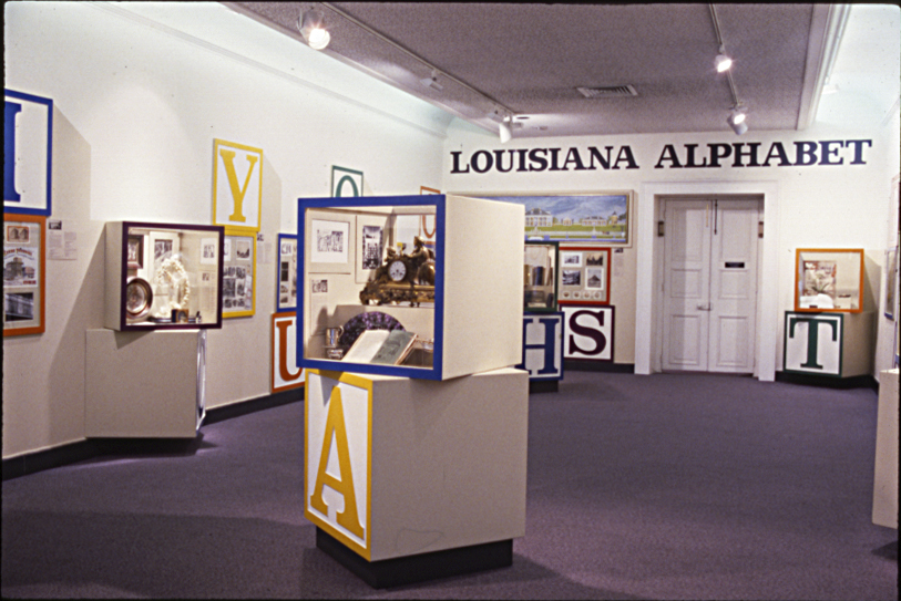 Photograph showing a gallery installation from the 1984 exhibition "Louisiana Alphabet." The name of the exhibition is written on a wall at the back of the room, with building-block style displays filled with objects.
